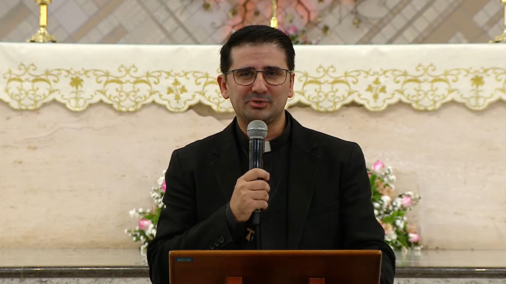 O padre de óculos e vestes pretas prega com um microfone no altar da igreja. Atrás dele, há um detalhado arranjo de flores e uma toalha litúrgica bordada.
