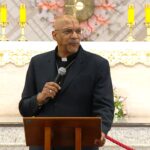 O sacerdote prega no altar do Santuário do Pai das Misericórdias, segurando um microfone atrás do ambão de madeira. Ao fundo, velas acesas ornamentam o presbitério durante a celebração litúrgica. Tema: "Quem não ama, não conhece a Deus"