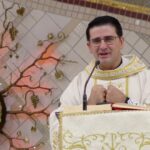 A imagem mostra um padre de óculos e vestes litúrgicas brancas e douradas discursando em um ambão decorado, com as mãos unidas junto ao peito. Ao fundo, destaca-se um sacrário circular cercado por ornamentos que simulam raízes e galhos de videira com cachos de uva sobre uma parede espelhada. Tema: "Santíssima Trindade e o chamado a identidade de filhos de Deus"