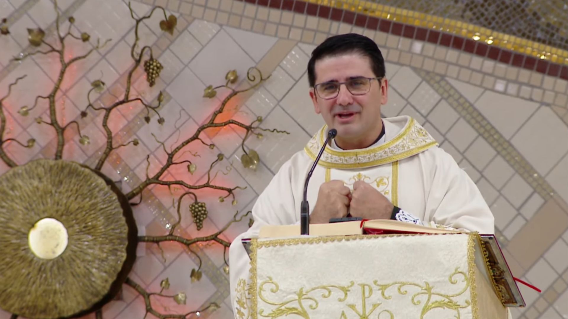 A imagem mostra um padre de óculos e vestes litúrgicas brancas e douradas discursando em um ambão decorado, com as mãos unidas junto ao peito.Ao fundo, destaca-se um sacrário circular cercado por ornamentos que simulam raízes e galhos de videira com cachos de uva sobre uma parede espelhada. Tema: "Santíssima Trindade e o chamado a identidade de filhos de Deus"