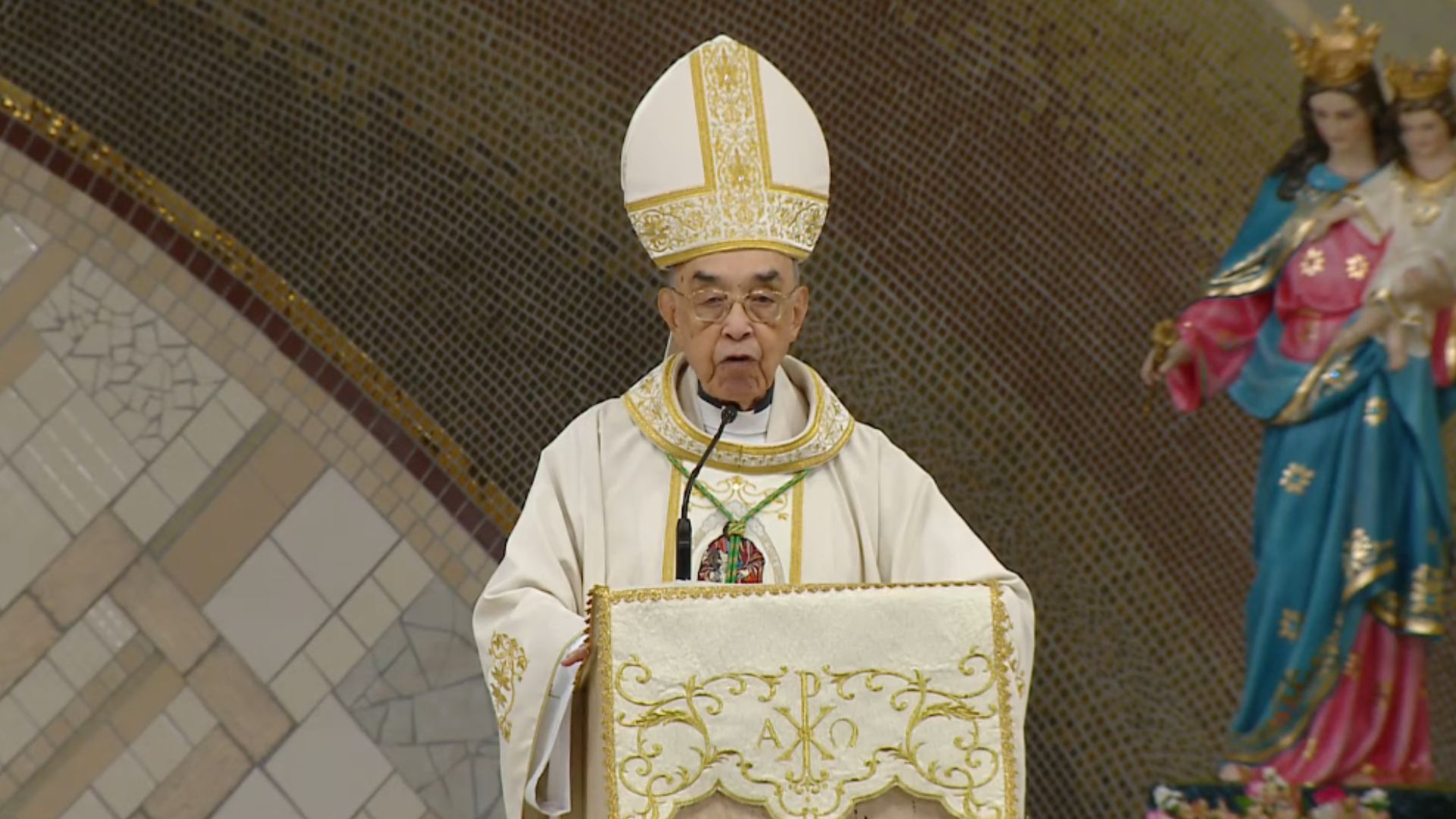 Bispo vestindo uma mitra e uma túnica brancas com detalhes dourados, discursa em um ambão decorado com símbolos cristãos. Ao fundo, uma parede de mosaicos dourados completa o cenário religioso ao lado de uma estátua de Nossa Senhora com o Menino Jesus. Tema: "Santíssima Trindade o caminho para a comunhão do amor"