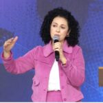 Mulher de cabelos pretos e cacheados, vestindo jaqueta e saia na cor rosa, fala ao microfone em cima de um palco. Tema: A “sincera doação de si”