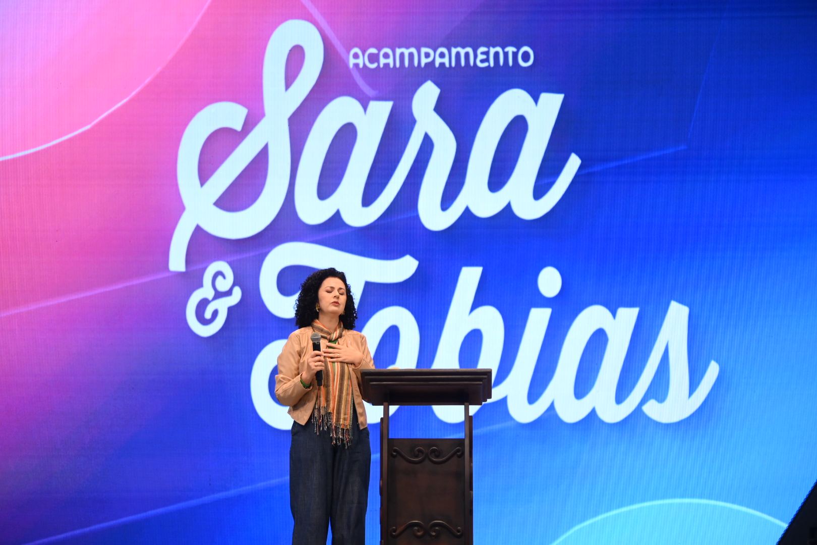Mulher de cabelos pretos e cacheados, vestindo jaqueta e saia na cor rosa, fala ao microfone em cima de um palco. Tema: A “sincera doação de si”