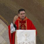 Padre de óculos e túnica vermelha gesticulando enquanto fala em um ambão com pano bordado. Ao fundo, destaca-se uma parede em arco revestida de mosaicos dourados e uma estátua de Nossa Senhora à direita. Descubra como o Espírito Santo podem renovar sua fé, dar coragem para enfrentar perseguições e transformar seu coração e gerando frutos na sua missão.