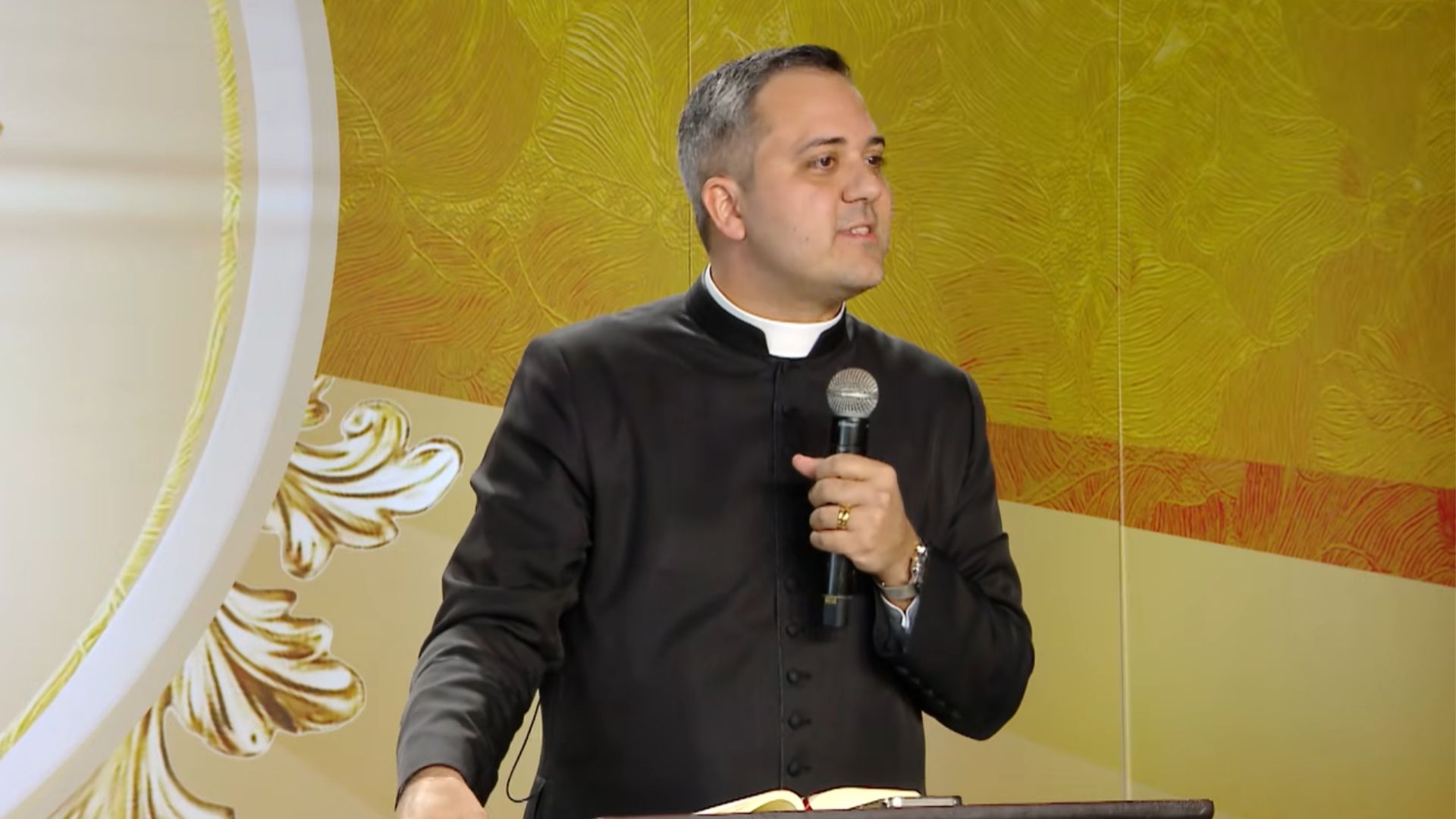 Um padre fala com um microfone na mão em um pódio, usando uma batina preta e colarinho clerical.O fundo é dourado com texturas e um detalhe floral.