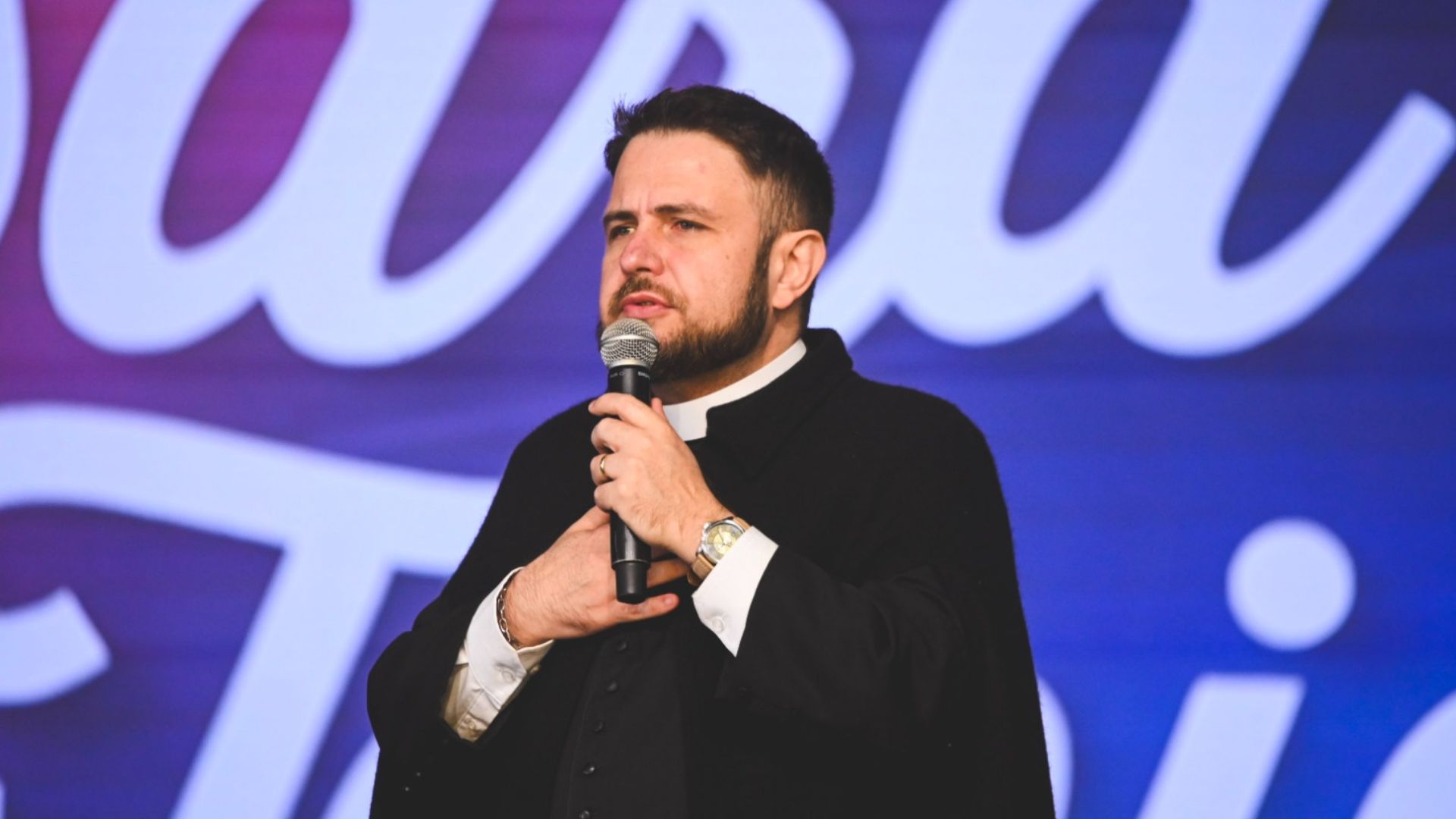 Sacerdote jovem com barba, usando vestes clericais pretas falando ao microfone enquanto gesticula com a mão no peito. Tema: "É na oração que se forja um homem valoroso"