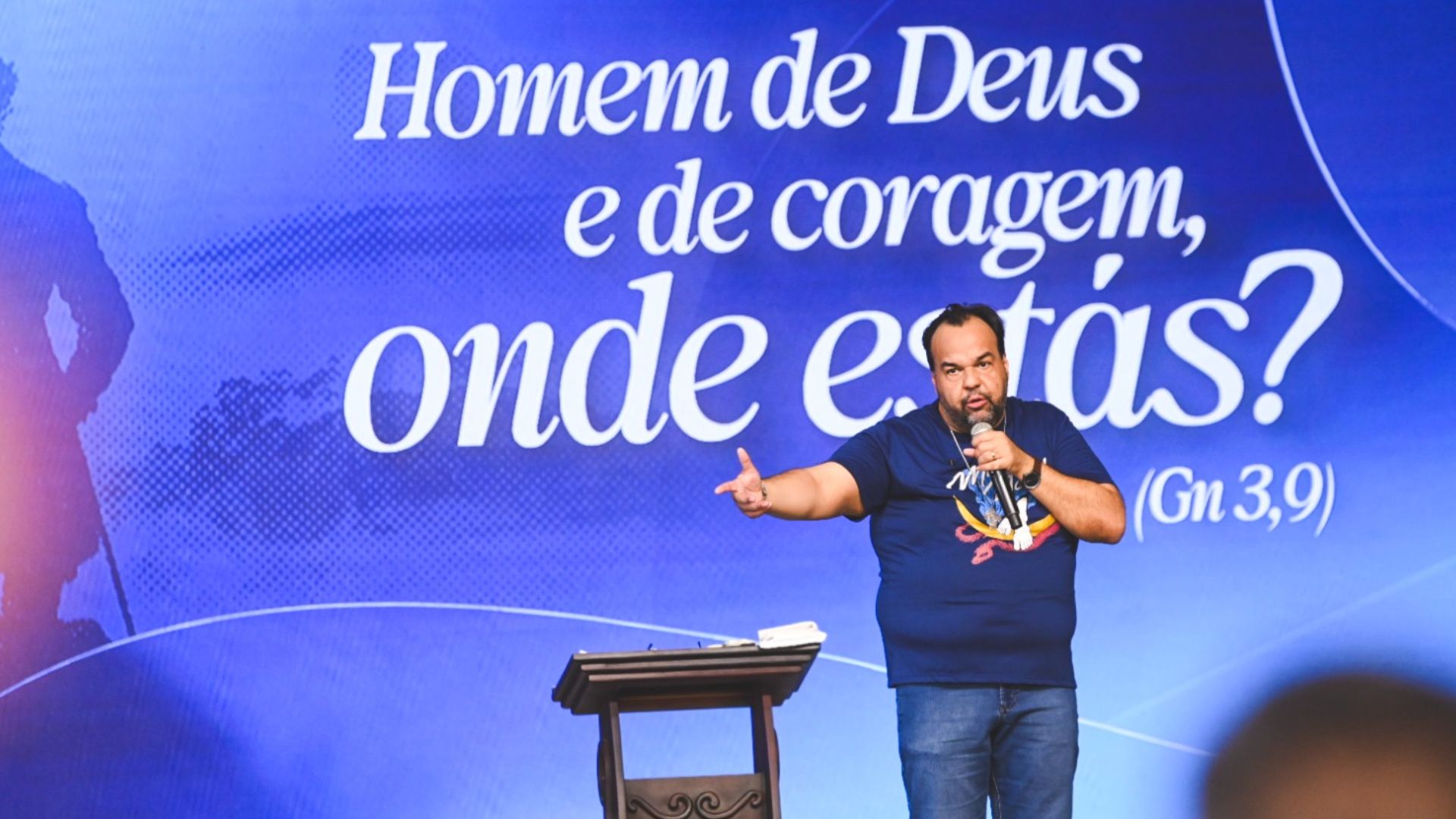 Homem, com uma camisa azul, de pé num palco a falar ao microfone. Atrás dele, há um painel azul com as palavras "Homem de Deus e de coragem, onde estás?"