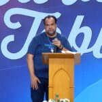 Um homem de camiseta azul fala ao microfone atrás de um púlpito de madeira claro. Tema: "Homem e Mulher em busca da unidade da alma e do corpo"