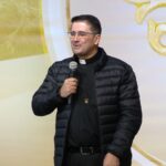 Descubra como a adoração a Jesus sacramentado, nosso Deus amado, transforma vidas. Inspire-se com testemunhos sobre o poder da Eucaristia. Acesse e leia!Ele segura um microfone com a mão direita e fala em um palco com fundo decorado em tons de dourado e branco, contendo detalhes religiosos.
