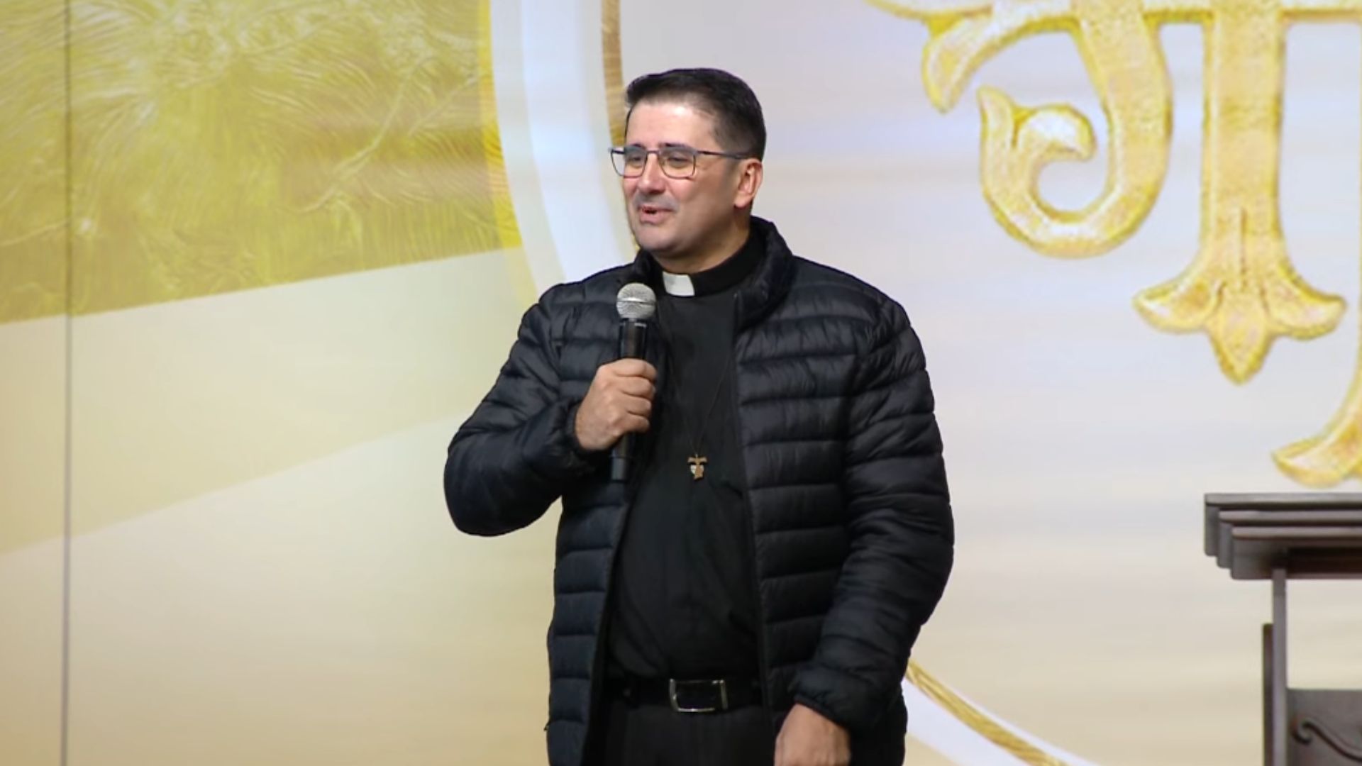 Padre sorridente usando óculos, camisa preta com colarinho clerical e uma jaqueta puffer preta aberta.Ele segura um microfone com a mão direita e fala em um palco com fundo decorado em tons de dourado e branco, contendo detalhes religiosos.