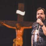 Homem falando ao microfone com uma expressão expressiva em um palco. Ao fundo, em um plano desfocado sob um fundo escuro, destaca-se uma grande escultura de Jesus Cristo crucificado. Tema: Maridos, amem as suas esposas!"
