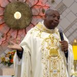 Padre vestindo uma túnica branca e dourada ornamentada, segurando um microfone no altar de uma igreja. Ao fundo, destaca-se um grande ostensório dourado na parede decorada, ladeado por velas amarelas acesas em castiçais dourados.