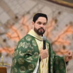 Um padre com barba e vestes litúrgicas verdes e douradas segura um microfone enquanto gesticula e prega em um altar. Ao fundo, destaca-se uma parede ornamentada com uma árvore decorativa iluminada em tons quentes. Tema: "O tesouro encontrado em um coração curado"