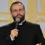Padre jovem de barba e óculos, vestindo uma batina preta com colarinho clerical branco, enquanto segura um microfone e fala. Ao fundo, destaca-se um painel decorativo em tons dourados e claros.