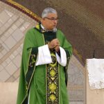 Padre de óculos e casula verde, segurando um microfone com as duas mãos durante uma pregação. Ele está no presbitério, ao lado de um ambão com uma toalha branca bordada. tema: "Santa Eucaristia o remédio que cura a alma"
