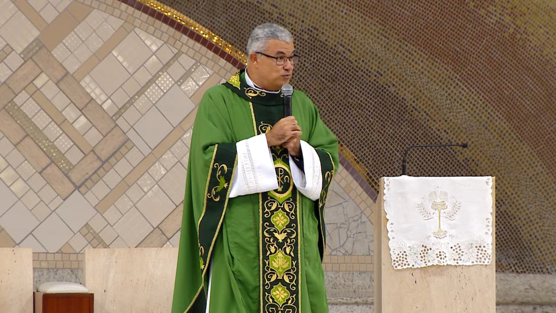 Padre de óculos e casula verde, segurando um microfone com as duas mãos durante uma pregação. Ele está no presbitério, ao lado de um ambão com uma toalha branca bordada. tema: "Santa Eucaristia o remédio que cura a alma"