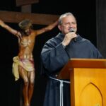 Um frei sorri ao falar ao microfone em um púlpito de madeira, vestindo seu tradicional hábito escuro com cordão branco. Ao fundo, destaca-se uma grande imagem esculpida de Jesus Cristo crucificado sobre um cenário escuro. Tema: "Sem fé é impossível agradar o coração de Deus"