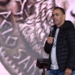 Um homem de jaqueta preta acolchoada fala ao microfone no que parece ser uma palestra ou pregação em um palco.