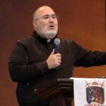 Sacerdote de terno preto sobre uma camisa preta, de óculos, e com a mão direita segurando um microfone.