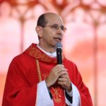 padre calvo com óculos de grau usando um paramento litúrgico vermelho brilhante enquanto fala ao microfone. O preço que Deus pagou para nos salvar revela o verdadeiro sentido da cruz. Compreenda como o sangue de Cristo nos fortalece diante das tribulações.