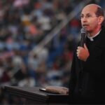 Um padre usando óculos e casaco preto fala ao microfone em cima de um palco com uma grande multidão desfocada ao fundo. Descubra como o "Cor Inquietum" de Santo Agostinho revela o caminho da conversão. Uma reflexão sobre o pecado, a graça e a decisão de ser santo hoje.