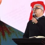 Padre de cabelos grisalhos e óculos, com uma blusa de gola alta preta, no púlpito, segurando um microfone e olhando para a direita. Tema: "Educai-nos para a santidade"
