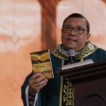 Um padre com óculos e vestes litúrgicas verdes fala ao microfone atrás de um púlpito de madeira. Ele segura e exibe o livro "A Bíblia no meu dia-a-dia", do Monsenhor Jonas Abib, contra um fundo em tons pastéis.