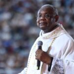Sacerdote vestindo túnica litúrgica branca com detalhes dourados, segura um microfone com expressão serena. Ele está de óculos e direciona seu olhar adiante, tendo ao fundo uma grande multidão desfocada.