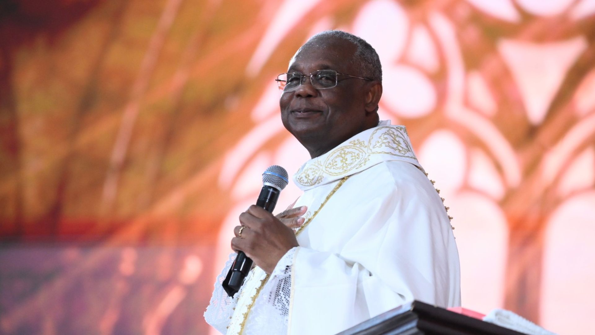 Padre sorridente de óculos. Ele veste uma túnica branca com detalhes dourados e segura um microfone com a mão direita, enquanto a esquerda está sobre o peito.