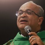 Sacerdote com veste litúrgica verde usando óculos e segurando um microfone.