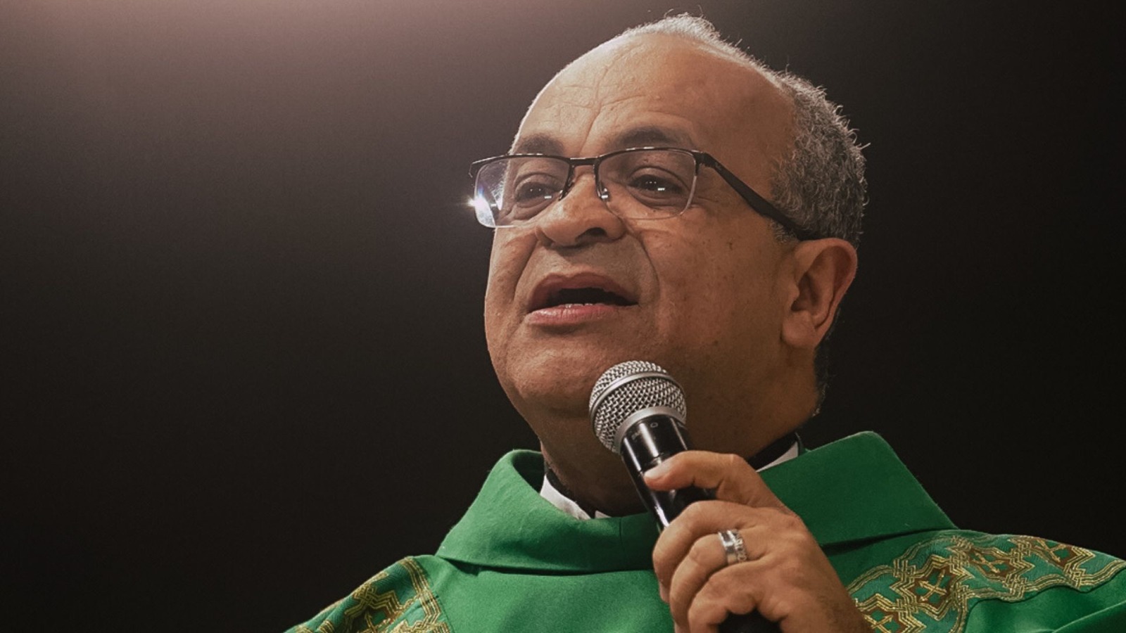 Sacerdote com veste litúrgica verde usando óculos e segurando um microfone.