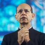 Um homem de óculos e veste clerical preta aparece em um momento de profunda devoção e reflexão. Com as mãos unidas em sinal de oração.