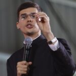 Jovem padre compartilha sua mensagem com expressividade enquanto gesticula firmemente com uma das mãos. Ele veste sua batina preta tradicional, usa óculos e segura com precisão o microfone próximo à boca durante a pregação.