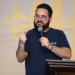 Um homem com barba cheia, óculos de grau e camiseta azul-marinho fala ao microfone atrás de um púlpito de madeira. Ele faz um gesto com o punho direito cerrado, diante de um fundo claro com detalhes religiosos amarelos. Tema: "Portanto, irmãos, cuidai cada vez mais em assegurar a vossa vocação e eleição"