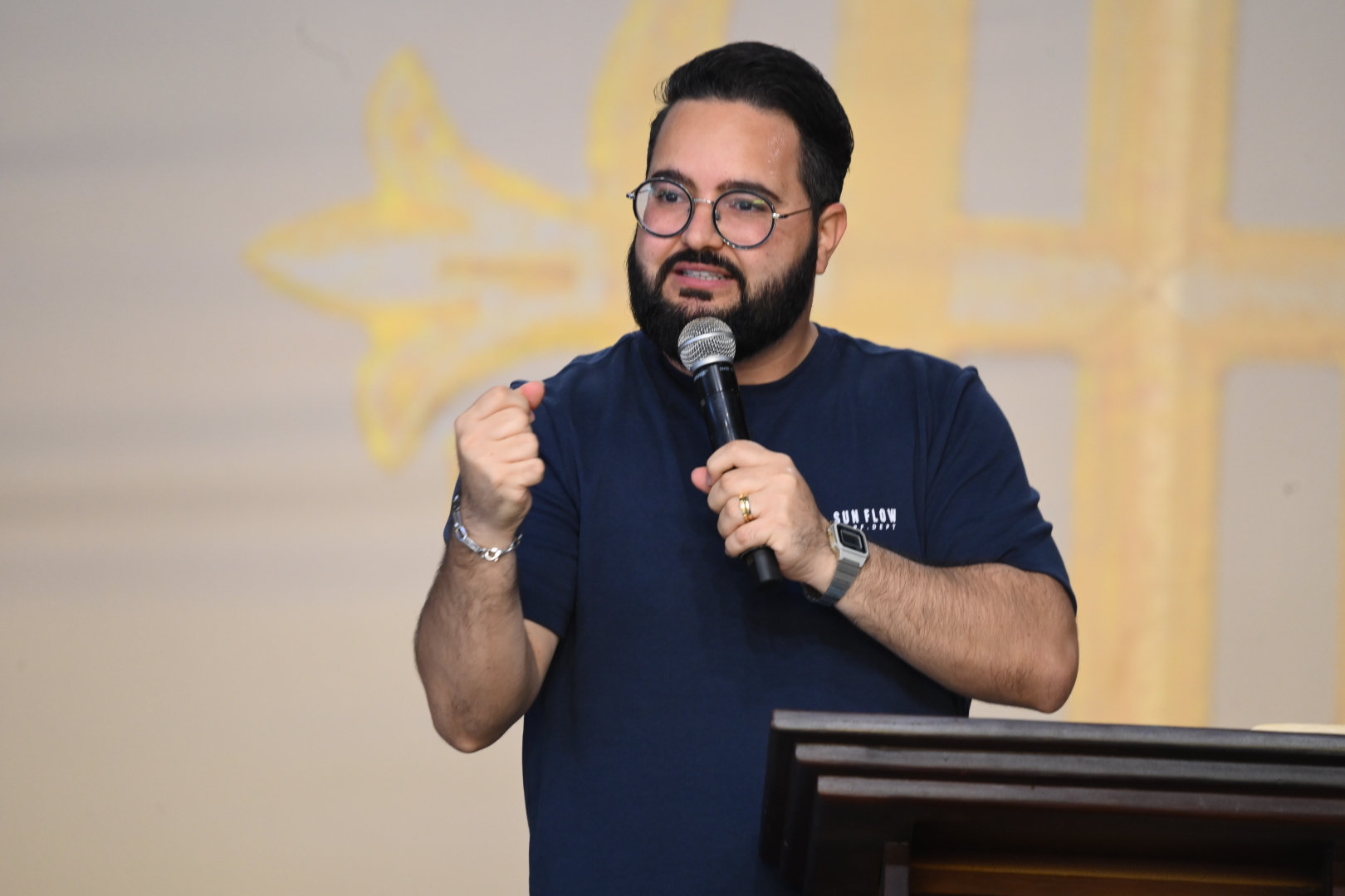 Um homem com barba cheia, óculos de grau e camiseta azul-marinho fala ao microfone atrás de um púlpito de madeira. Ele faz um gesto com o punho direito cerrado, diante de um fundo claro com detalhes religiosos amarelos.Tema: "Portanto, irmãos, cuidai cada vez mais em assegurar a vossa vocação e eleição"