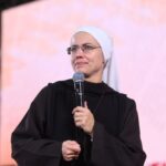 Uma religiosa de óculos e hábito escuro gesticula com uma das mãos enquanto fala ao microfone no palco.