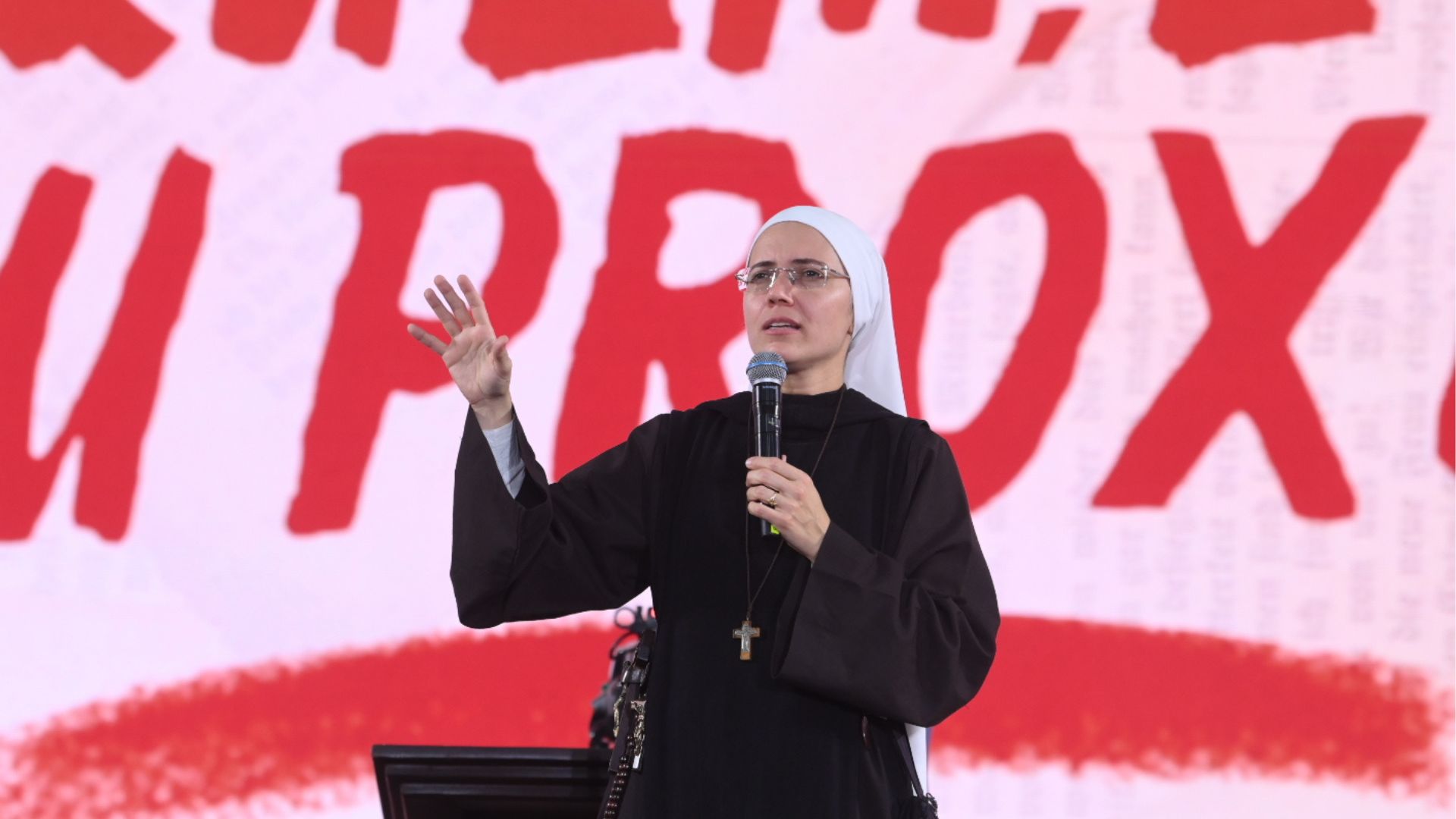 Uma religiosa de óculos e hábito escuro gesticula com uma das mãos enquanto fala ao microfone no palco.Ao fundo, destaca-se um grande painel com letras vermelhas garrafais que trazem a frase "QUEM É O MEU PRÓXIMO".