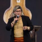 Uma mulher fala ao microfone segurando um rosário. Ela veste uma blusa bege, calças cor de terracota e um casaco preto