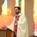 Um padre jovem com barba usando vestes litúrgicas douradas e vermelhas fala ao microfone em um púlpito de madeira. Ele gesticula com a mão direita em frente a vitrais coloridos. Tema: "A ação de Deus e os guarda-chuvas da alma"