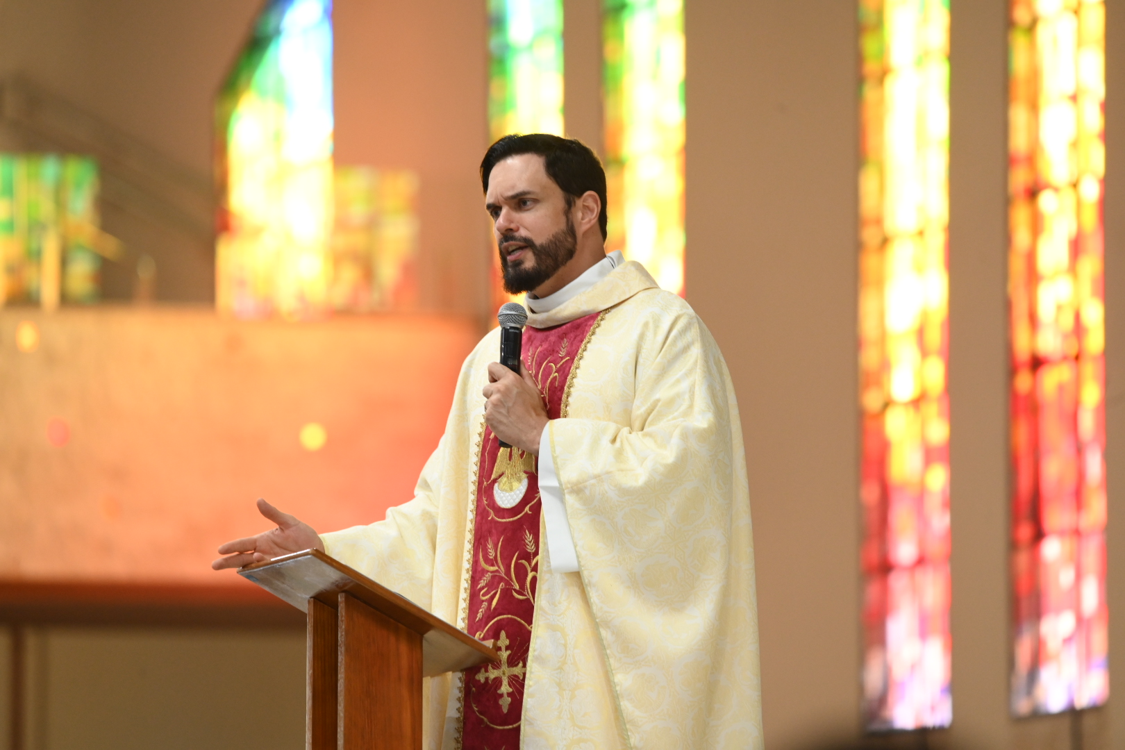 Um padre jovem com barba usando vestes litúrgicas douradas e vermelhas fala ao microfone em um púlpito de madeira. Ele gesticula com a mão direita em frente a vitrais coloridos. Tema: "A ação de Deus e os guarda-chuvas da alma"