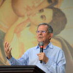 Professor Felipe Aquino fala com entusiasmo em um microfone atrás de um pódio de madeira. O fundo desfocado da pintura exibe uma representação da Virgem Maria e do menino Jesus. Tema: A continuidade do poder de Cristo através dos Sacramentos!"