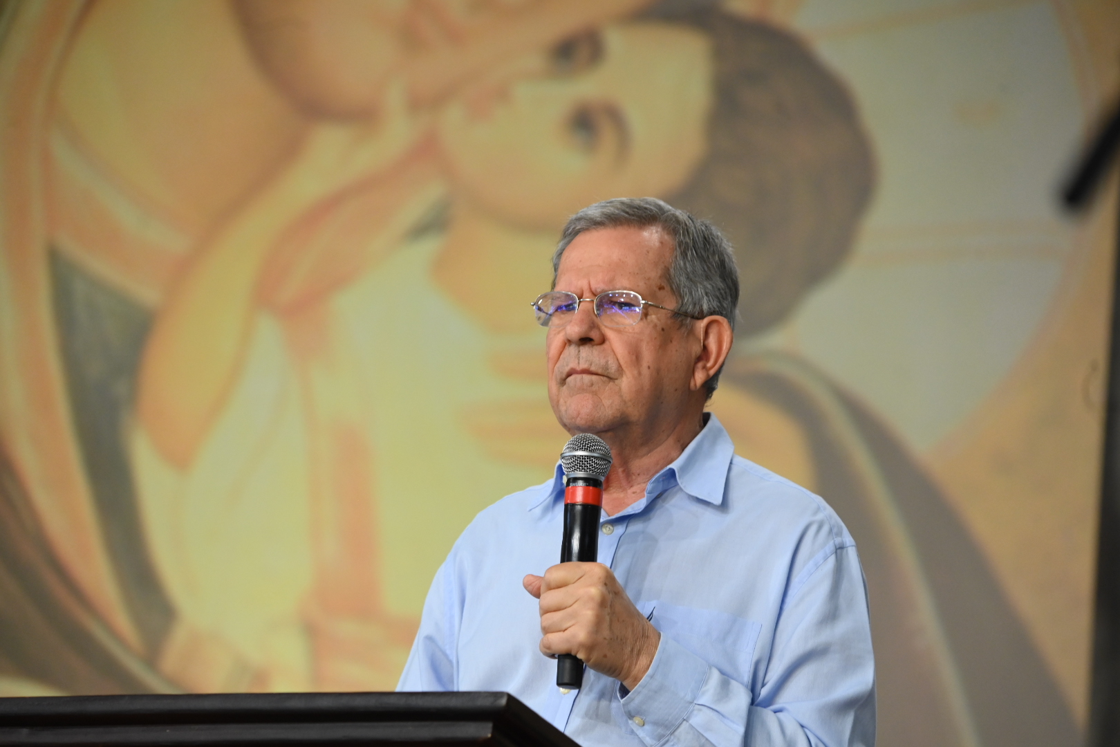 Professor Felipe Aquino segura um microfone com expressão serena e atenta atrás de um pódio.Ao fundo, destaca-se a imagem em tom suave de um ícone sacra representando a Virgem Maria com o Menino Jesus.