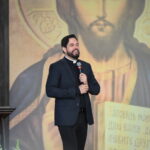 Um padre jovem de barba, vestindo terno escuro e clergyman sorri enquanto fala ao microfone. Ao fundo, há um grande painel com a imagem sacra de Jesus Cristo segurando as Escrituras. Tema: "A fidelidade do cristão em tempos de relativismo!"