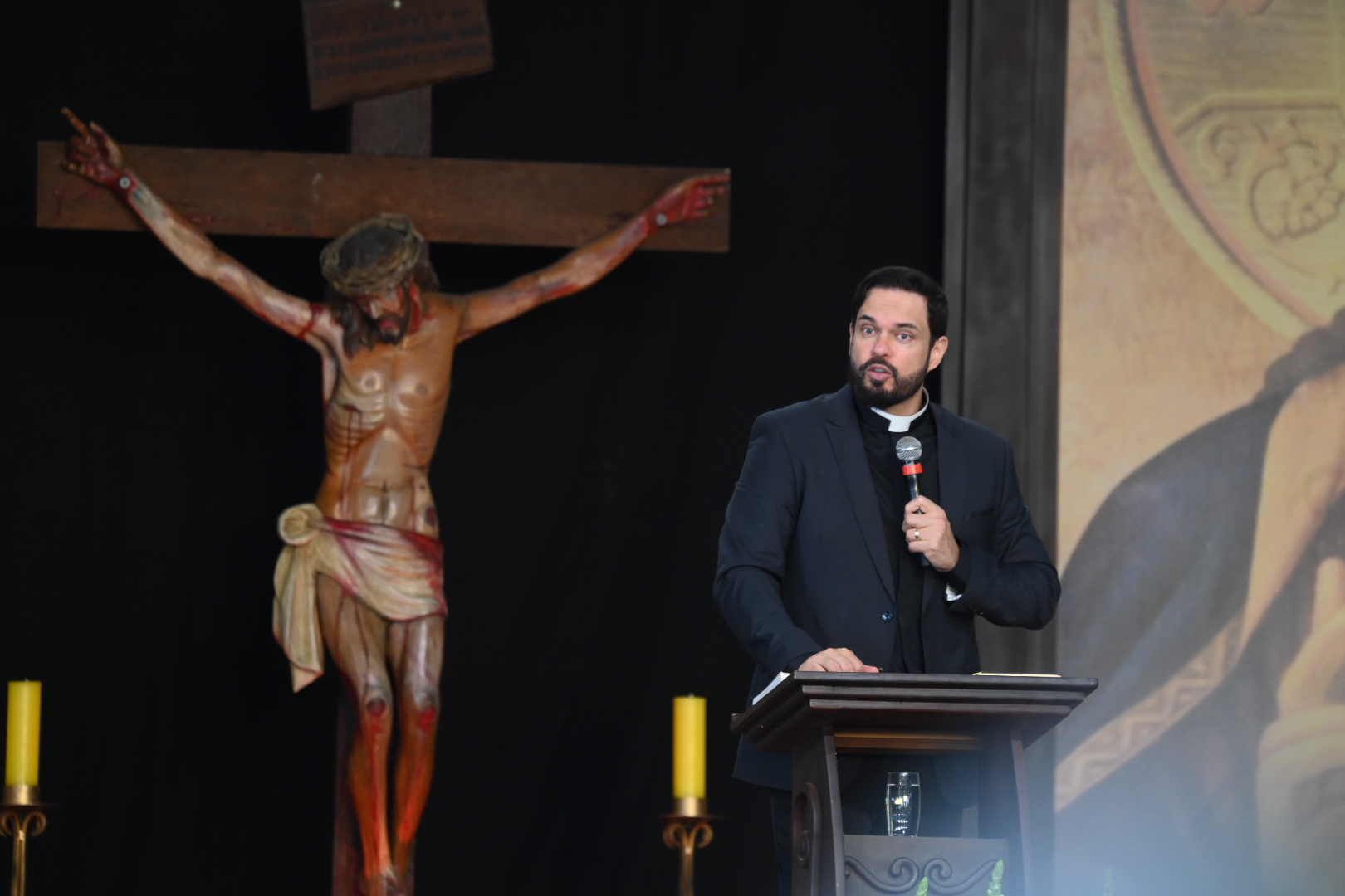 Um padre jovem de barba, vestindo terno escuro e clergyman fala ao microfone em um púlpito de madeira.Ao lado esquerdo, em segundo plano, destaca-se uma grande escultura de Jesus Crucificado.