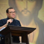 Um padre de óculos e camisa clerical escura fala ao microfone apoiado em um púlpito de madeira. Tema: "Um padre de óculos e camisa clerical escura fala ao microfone apoiado em um púlpito de madeira."