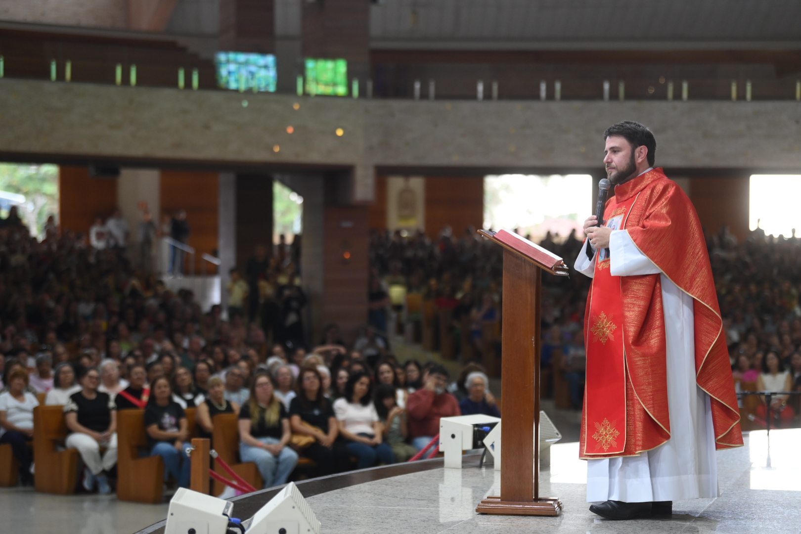 Um padre vestindo vestes litúrgicas vermelhas e brancas fala ao microfone ao lado de um ambão de madeira em um altar elevado.Ao fundo, uma grande assembleia de fiéis acompanha a celebração no interior de uma igreja ampla. Tema: "Acalmou-se a Vossa ira e enfim me consolastes"