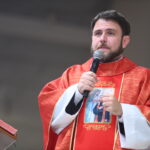Um padre vestindo vestes litúrgicas vermelhas fala ao microfone em um púlpito, gesticulando com a mão esquerda enquanto segura o microfone com a direita. Tema: "Acalmou-se a Vossa ira e enfim me consolastes"