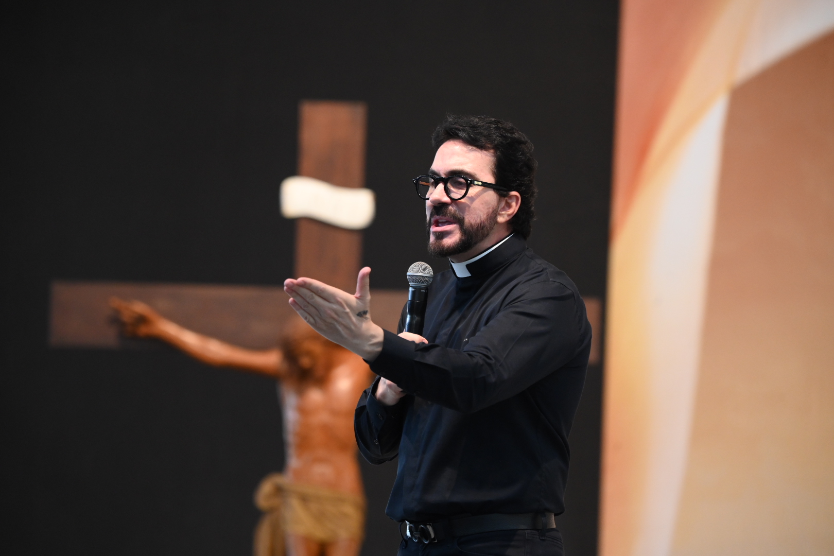 Fábio de Melo aparece de perfil em um altar, vestindo camisa clerical preta e óculos, enquanto segura um microfone e gesticula com a mão aberta.Ao fundo, desfocado, vê-se um crucifixo de madeira em destaque sobre um painel escuro. Tema: "As febres que nos privam de conhecer a verdade"