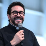 Padre Fábio de Melo aparece sorrindo enquanto segura um microfone e fala ao público. Tema: "As febres que nos privam de conhecer a verdade"