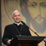 Um bispo católico sorridente, com veste preta, cruz peitoral e solidéu, fala ao microfone em um púlpito de madeira.